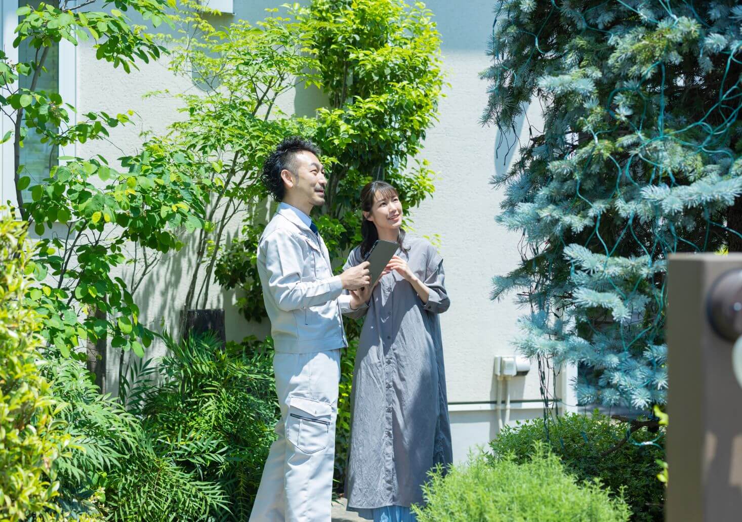 緑に囲まれた庭先で、作業着の担当者がタブレットを見せながらご婦人と話している様子