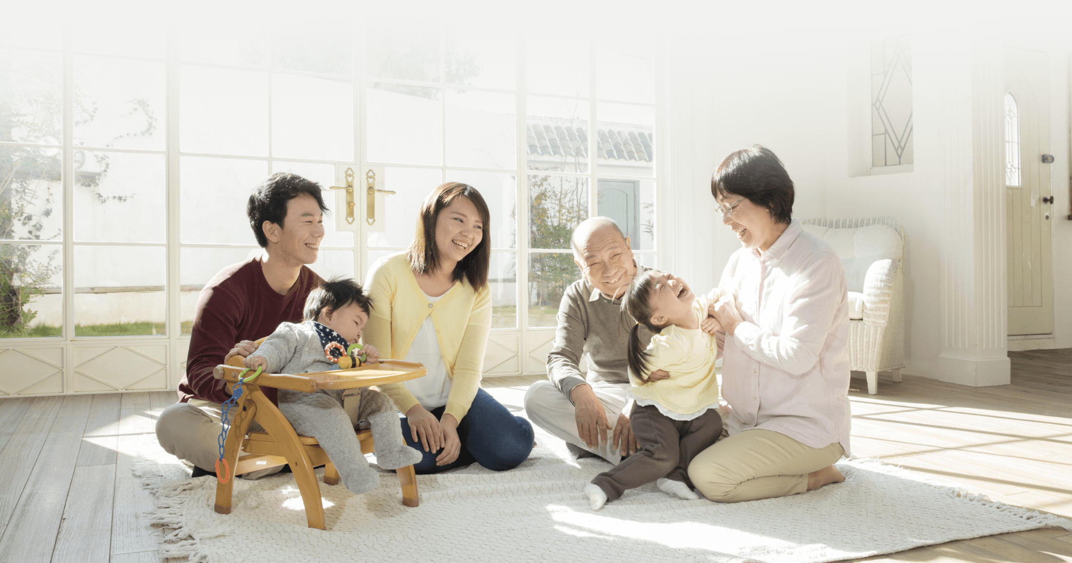 大きな窓から日の光が差し込む明るいリビングで、祖父母から赤ちゃんまで三世代家族が笑顔で過ごす様子